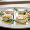 Marinated goat cheese medallions drizzled with herb-infused olive oil and fresh rosemary on white plate