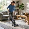 A person using an anti-allergen vacuum on a carpet with a dog sitting nearby in a bright living room