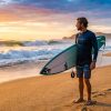 Surfer in high-performance rash guard waxing a surfboard on a sunny beach.